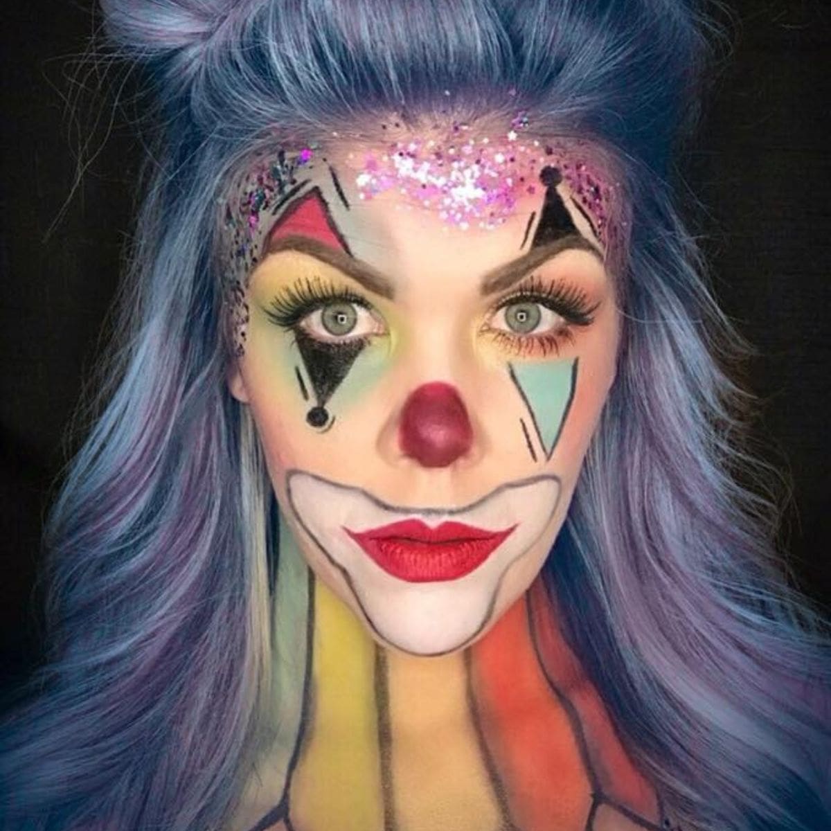 Person with light purple hair styled up, wearing colorful clown makeup with bold shapes in red, yellow, blue, and black on the face and neck. Glitter detail on forehead and bright red lips. Black background.