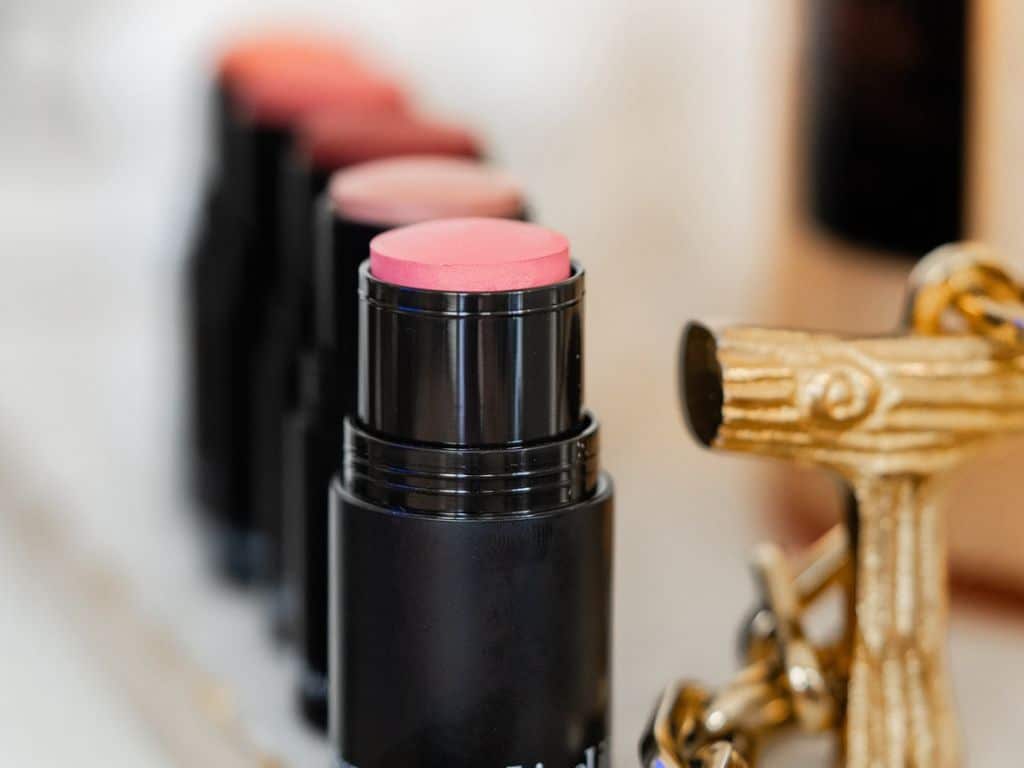 A close-up of several stick blushes with pink and coral tones in black tubes, arranged in a row. A gold-colored keychain shaped like a T-bar is visible on the right side of the image.