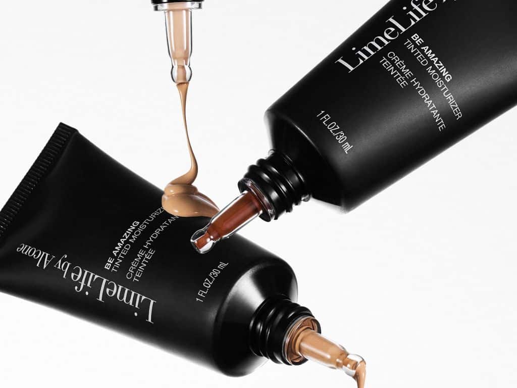 Three black tubes of LimeLife by Alcone tinted moisturizer with beige product being dispensed from two tubes and drops forming at the openings, set against a white background.